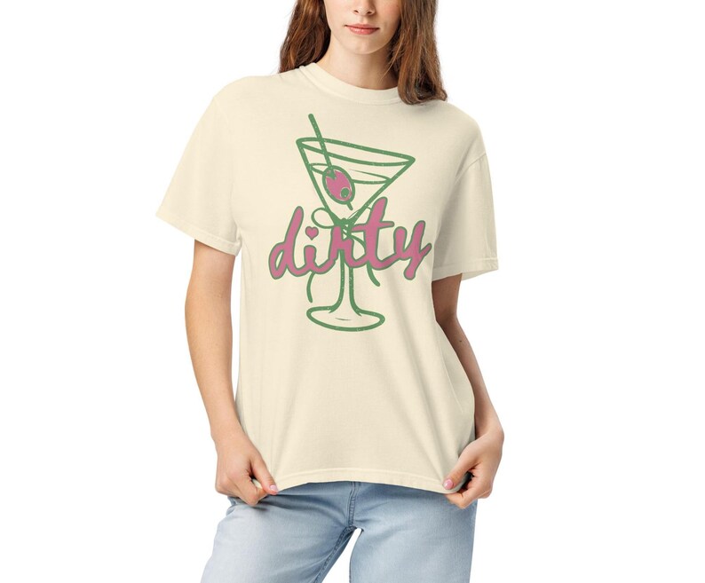 May include: Cream-colored t-shirt featuring a green outline of a martini glass with an olive and a straw. The word "dirty" is written in pink script below the glass. The shirt is made of a soft material.