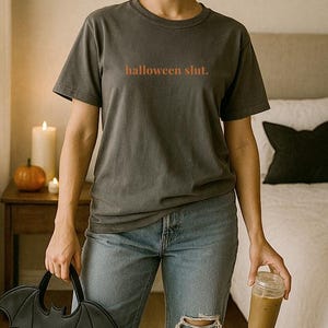 May include: Dark gray t-shirt with the text "halloween slut." in orange. The person is wearing blue jeans and holding a black bat-shaped handbag and a clear glass with a brown beverage.