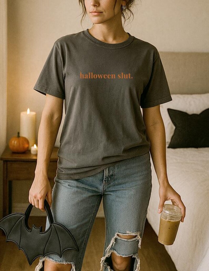 May include: Dark gray t-shirt with the text "halloween slut." in orange. The person is wearing blue jeans and holding a black bat-shaped handbag and a clear glass with a brown beverage.