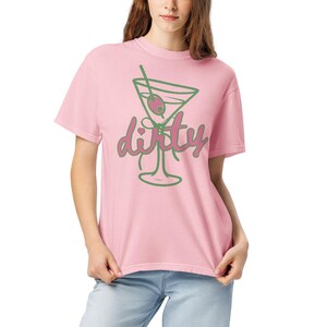 May include: A light pink t-shirt featuring a green outline of a martini glass with an olive and a straw. The word "dirty" is written in a cursive font in pink and green. The shirt is made of a soft material.