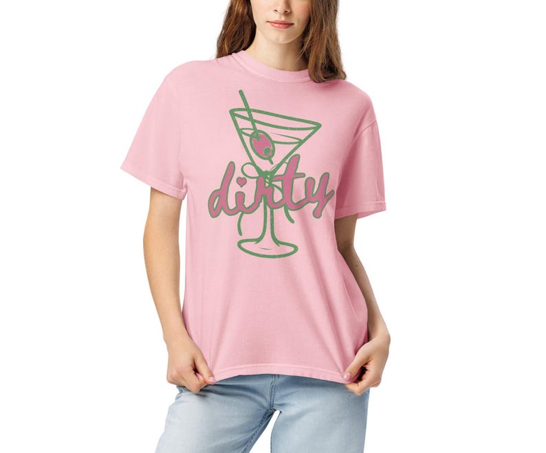 May include: A light pink t-shirt featuring a green outline of a martini glass with an olive and a straw. The word "dirty" is written in a cursive font in pink and green. The shirt is made of a soft material.