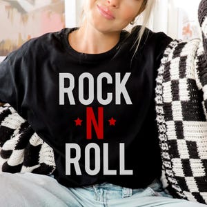 May include: Black t-shirt with white text that reads "ROCK N ROLL" with a red "N" and white stars on either side of the "N".
