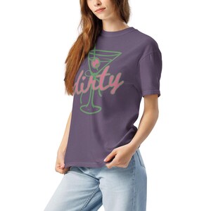 May include: A purple t-shirt featuring a green and pink martini glass design with the word "dirty" in a cursive font. The shirt is made of a soft material and is being worn by a person.