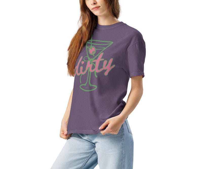 May include: A purple t-shirt featuring a green and pink martini glass design with the word "dirty" in a cursive font. The shirt is made of a soft material and is being worn by a person.