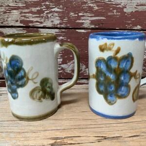 May include: Two ceramic mugs with a grapevine design. One mug has a brown rim and the other has a blue rim.