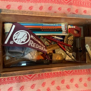 May include: An open wooden drawer filled with various items, including a burgundy pennant with the text "WASHINGTON REDSKINS", pencils, pens, a glass jar, and a small wooden box.