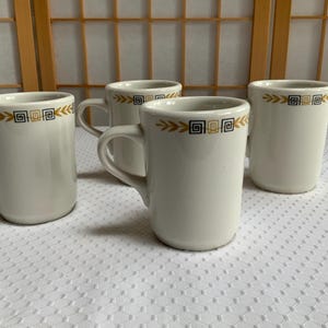 May include: Four white ceramic mugs with a gold and black geometric design around the rim. The mugs have a simple, classic shape with a handle on the side. They are arranged on a white textured surface.