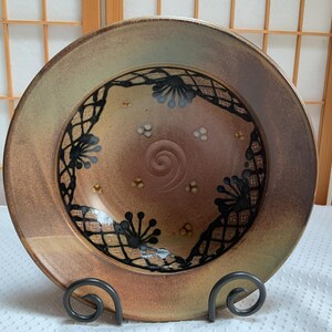 May include: A round ceramic bowl with a wide rim, featuring a metallic bronze and copper glaze. The interior displays a black lace-like pattern with floral motifs, small white dot accents, and a central spiral indentation in the clay surface.