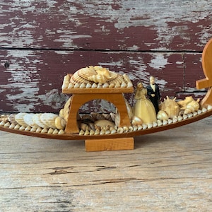 May include: A wooden gondola decorated with seashells and featuring a bride and groom figurine. The gondola is a wedding keepsake or decoration.