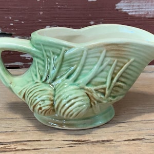 May include: A vintage, light green ceramic creamer with a handle and a spout. The creamer is decorated with a relief pattern of leaves and bulbous shapes. The base is a light beige color.