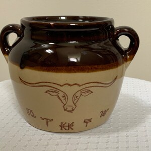 May include: Brown and tan ceramic crock with two handles. The crock has a brown glaze and a design of a bull's head with horns and the text "B.T. Kraft W."