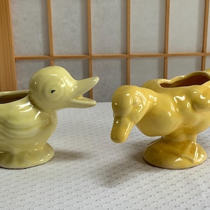 May include: Two ceramic duck planters in a light yellow color. One duck is upright with its beak open, and the other is in a downward position. Both have a hollowed-out area for planting.