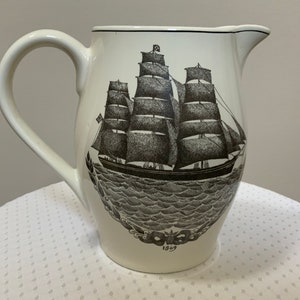 May include: White ceramic pitcher with a black and white illustration of a sailing ship with the year 1869 printed on the bottom.