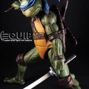 May include: A Teenage Mutant Ninja Turtle action figure, green with a blue mask, holding two katana swords. The figure is posed in a dynamic stance, with a yellow plastron and brown straps. The text "EQUIP custom action figure mini weapons" is visible.