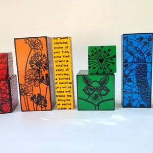 May include: A set of colorful wooden blocks with different designs. The blocks are stacked in a rainbow pattern, with each block featuring a unique image. The designs include flowers, butterflies, a peacock, a dog wearing glasses, and a heart. The blocks are made of wood and have a smooth, polished finish.
