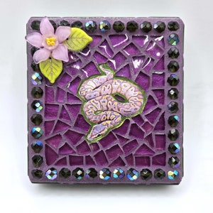 May include: A purple mosaic tile featuring a snake design and a pink flower with green leaves. The tile is bordered by black iridescent beads.