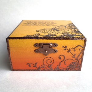 Puede incluir: Una pequeña caja de madera con un acabado de pintura degradado amarillo y naranja. La caja tiene un diseño de mariposa y una cita que dice "Lo que haces en el presente es siempre el coraje para cambiar, la sabiduría para conocer la diferencia".