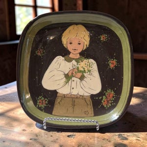 May include: A square ceramic plate with rounded corners featuring a central illustration of a person with blonde hair wearing a white long-sleeved shirt and tan pants, holding a bouquet of green and orange flowers against a dark brown background.