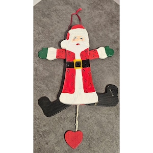 May include: A wooden Santa Claus Christmas ornament with outstretched arms and dangling legs. The ornament features a red coat with white trim, a black belt, and green mittens. A red heart hangs below.