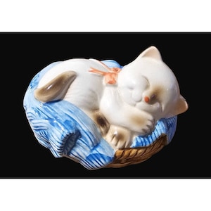 May include: A white ceramic figurine of a sleeping cat with a brown tail, lying on a blue and white striped bed with a brown wicker base.