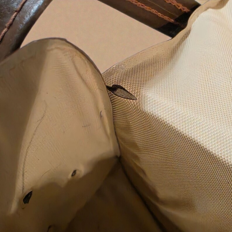 Puede incluir: Vista interior de un bolso de cuero marr&oacute;n. El forro interior es de color beige claro y la estructura del bolso es visible. La cremallera del bolso est&aacute; cerrada. El asa del bolso es visible en la parte superior de la imagen.