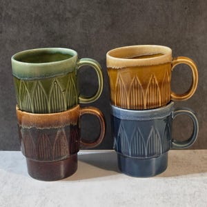 May include: A set of four ceramic mugs stacked in two pairs. Each mug features a vertical ribbed pattern, a smooth rim, and a glossy glaze. The colors include sage green, golden yellow, dark brown, and slate blue, with tonal variations.