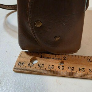 Puede incluir: Una funda de cuero marr&oacute;n con cierre de solapa y dos tachuelas color lat&oacute;n. Una regla de madera con marcas en cent&iacute;metros se coloca junto a la funda. La funda mide aproximadamente 7,6 cm de altura.