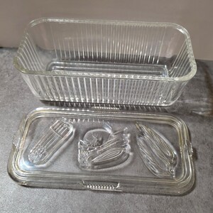 May include: A rectangular clear glass butter dish with a ribbed base and a matching lid. The lid features embossed designs of three distinct vegetable groupings, including asparagus stalks, a round vegetable, and long bean-like pods.
