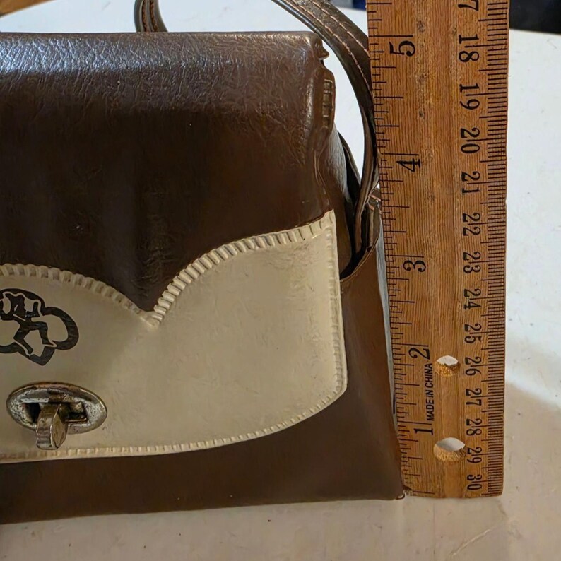 Puede incluir: Bolso vintage marr&oacute;n y crema con cierre plateado. El bolso presenta un dise&ntilde;o decorativo en la solapa delantera. La regla muestra una altura de aproximadamente 12,7 cm.