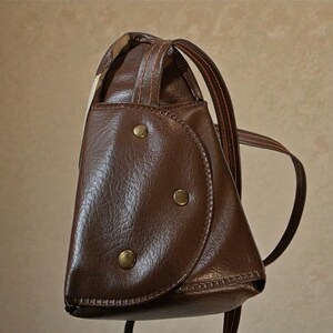 Puede incluir: Una mochila de cuero marr&oacute;n con una solapa redondeada asegurada por tres botones color lat&oacute;n. La bolsa tiene un asa superior y correas ajustables. La costura es visible y el dise&ntilde;o est&aacute; inspirado en lo vintage.
