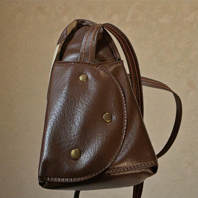 Puede incluir: Una mochila de cuero marr&oacute;n con una solapa redondeada asegurada por tres botones color lat&oacute;n. La bolsa tiene un asa superior y correas ajustables. La costura es visible y el dise&ntilde;o est&aacute; inspirado en lo vintage.