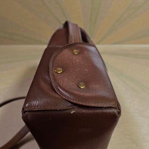 Puede incluir: Un bolso de mano de cuero marr&oacute;n con un asa superior y una correa para el hombro. El bolso tiene forma rectangular con bordes redondeados y est&aacute; asegurado con tachuelas de color lat&oacute;n. El dise&ntilde;o del bolso es vintage y cl&aacute;sico.