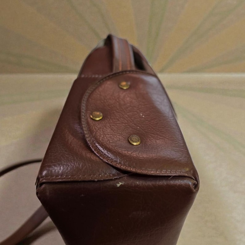 Puede incluir: Un bolso de mano de cuero marr&oacute;n con un asa superior y una correa para el hombro. El bolso tiene forma rectangular con bordes redondeados y est&aacute; asegurado con tachuelas de color lat&oacute;n. El dise&ntilde;o del bolso es vintage y cl&aacute;sico.