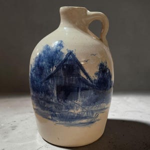 May include: A cream-colored ceramic jug featuring a blue hand-painted scene of a barn surrounded by trees. The vessel has a rounded body, a narrow neck, and a single handle attached to the side. The blue paint shows brushstroke textures.