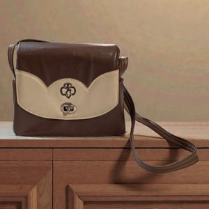 Puede incluir: Bolso de hombro marr&oacute;n y crema con cierre de solapa. El bolso presenta un emblema decorativo en la solapa delantera y un cierre plateado. La correa es de color marr&oacute;n.