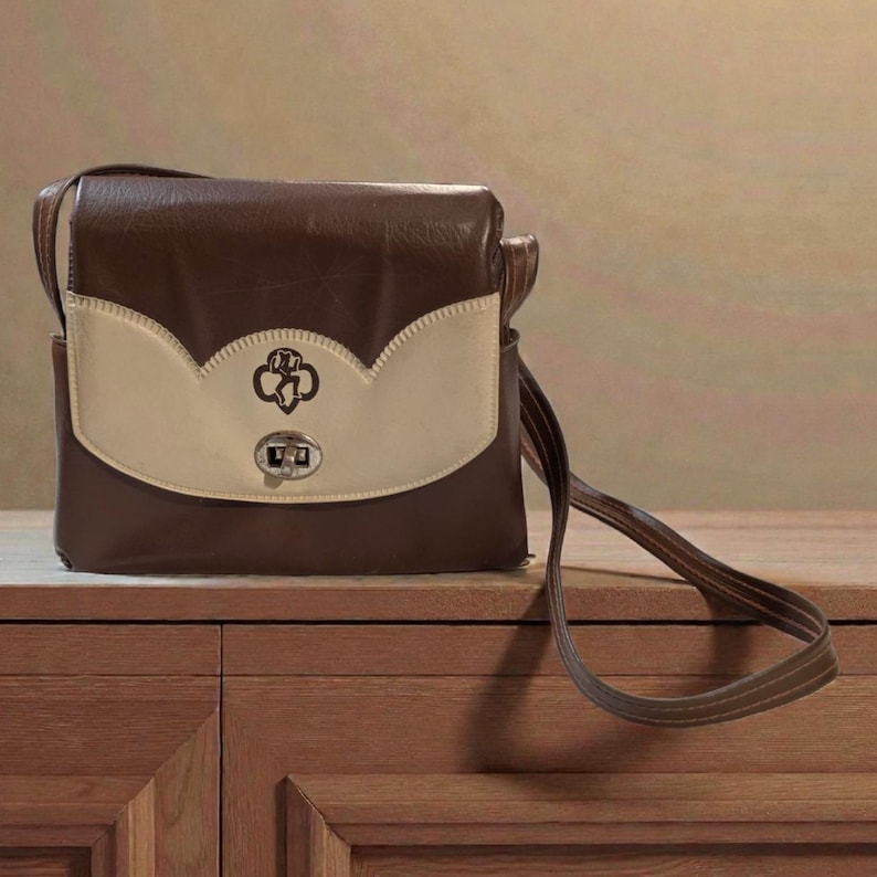 Puede incluir: Bolso de hombro marr&oacute;n y crema con cierre de solapa. El bolso presenta un emblema decorativo en la solapa delantera y un cierre plateado. La correa es de color marr&oacute;n.