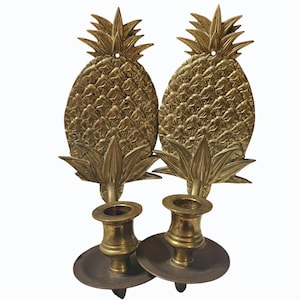 May include: Two brass pineapple-shaped wall sconces. Each sconce features a detailed pineapple design with a textured surface and a candle holder. The sconces are a warm gold color and are designed to be mounted on a wall.