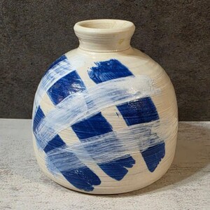 May include: A round ceramic vase with a cream-colored base featuring thick, overlapping brushstrokes in deep blue and white paint. The vase has a narrow neck with a flared rim and a textured surface showing horizontal ridges from the pottery process.
