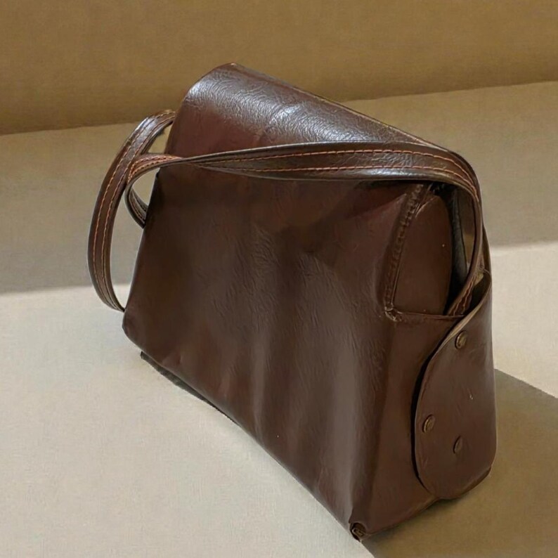 Puede incluir: Bolso de hombro de cuero marr&oacute;n con forma rectangular y parte superior curvada. El bolso tiene una correa larga y un cierre de solapa en el lateral. El bolso est&aacute; hecho de cuero liso y brillante.