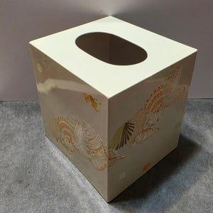 May include: A rectangular off-white tissue box cover featuring a decorative pattern of various seashells in shades of orange, brown, and green. The top surface has a large oval opening for dispensing tissues, and the sides display a repeating shell motif.