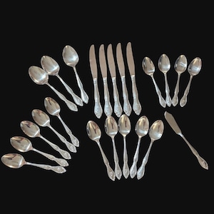 May include: A collection of silver-colored flatware, including spoons and knives, arranged on a black surface. The spoons have a rounded bowl and decorative handles. The knives have a smooth blade and a detailed handle design. A butter knife is also included.