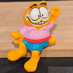 May include: A figurine of Garfield the cat, in a standing pose with one arm raised. The orange cat wears a pink shirt, blue shorts, and a red trim. The figurine has large white eyes and a smiling expression.