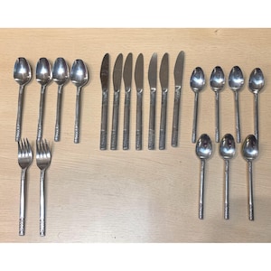 May include: A collection of stainless steel silverware, including spoons, forks, and knives. The spoons and forks have a textured handle design, while the knives have a smooth, simple design. The silverware is arranged on a light-colored wooden surface.