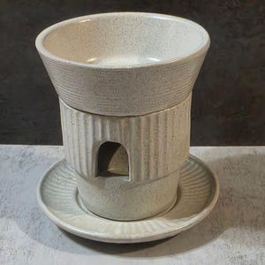 May include: A cream-colored ceramic oil warmer with a textured, ribbed design. The warmer has a bowl-shaped top, a cylindrical base with an arched opening, and a round base plate. The design is simple and elegant.