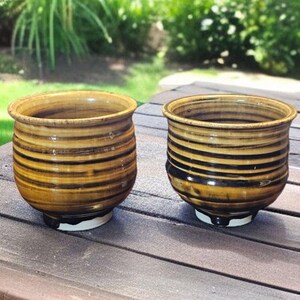 May include: Two handmade ceramic pots with a striped design in shades of brown and black. The pots have a rounded shape with a slightly flared rim and sit on a white base. The pots are displayed on a wooden surface.
