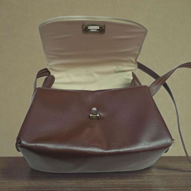 Puede incluir: Un bolso marr&oacute;n con forma rectangular y cierre de solapa. El bolso tiene una correa para el hombro y un interior de color claro. El bolso est&aacute; hecho de un material liso y tiene un cierre met&aacute;lico.