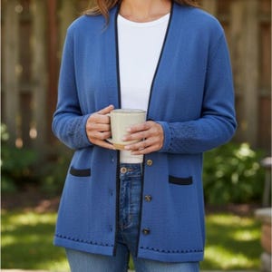 May include: A blue cardigan with black trim and button closures. The cardigan is worn over a white shirt and blue jeans. The sleeves have a decorative knit pattern. A beige mug is held in the hands.