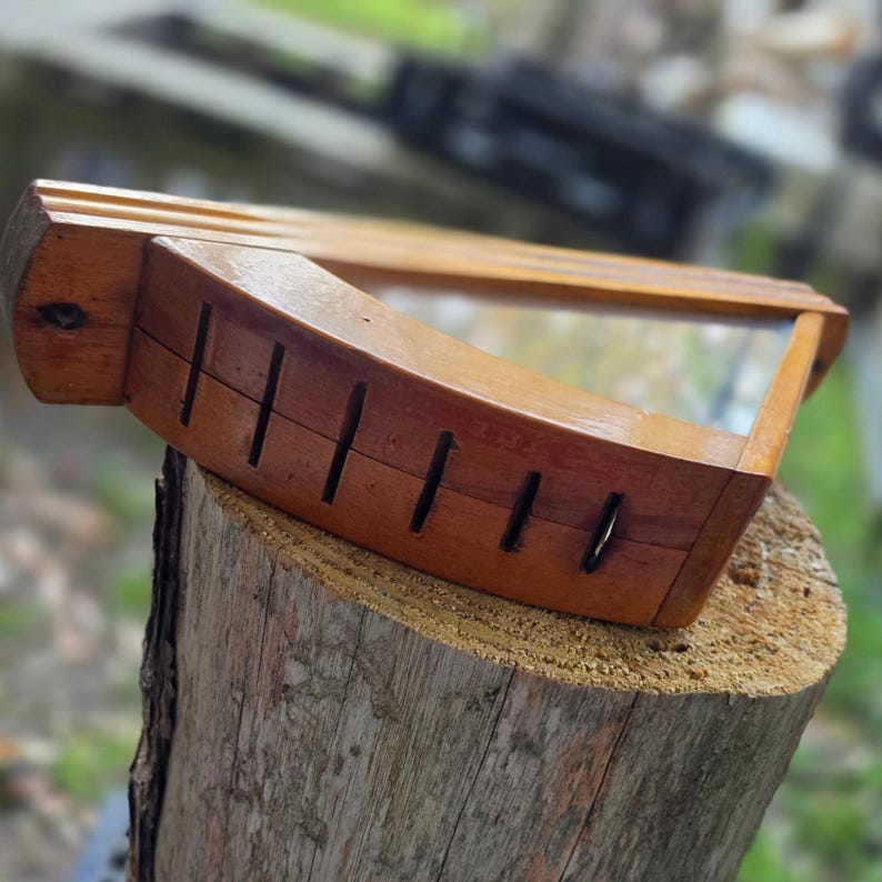May include: A wooden picture frame with a curved front and a rectangular glass pane. The frame is a warm brown color and has several vertical slots carved into the curved section. The frame is sitting on a tree stump.