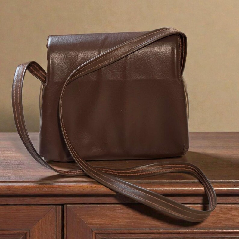 Puede incluir: Un bolso bandolera de cuero marr&oacute;n con cierre de solapa y una correa larga y ajustable. El bolso es rectangular con esquinas redondeadas y un ribete cosido. La correa tambi&eacute;n es marr&oacute;n y parece del mismo material que el bolso.