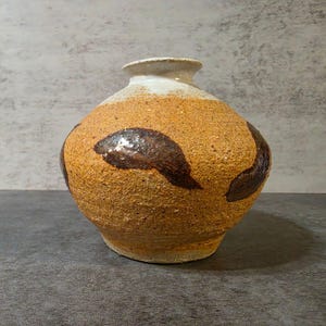 May include: A round ceramic vase with a textured, sandy tan body featuring dark brown, glossy, abstract brushstroke patterns. The neck and rim are finished in a smooth, off-white glaze that contrasts with the rough, earthy texture of the base.